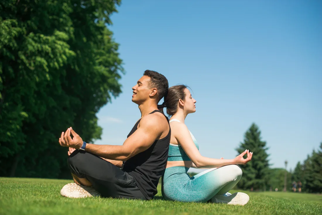 A man and a woman sitting back-to-back in meditation pose on grass in a park under a blue sky, set in a white frame